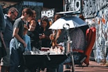 A group of people gathered around an outdoor chess game. A person is seated in front of a chessboard on the table, under a sunshade, with others standing and observing. The setting is urban, with graffiti-covered walls in the background.