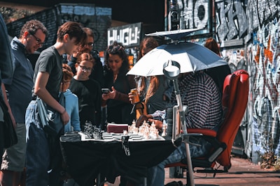 A group of people gathered around an outdoor chess game. A person is seated in front of a chessboard on the table, under a sunshade, with others standing and observing. The setting is urban, with graffiti-covered walls in the background.