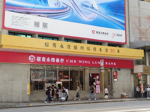 A bank building with signage for CMB Wing Lung Bank, featuring a large red and yellow sign with Chinese and English text. Several people are walking on the sidewalk in front of the building, and there are decorative stone lions by the entrance. Above the bank is a large blue and white advertisement billboard with cityscape graphics.