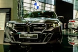 A sleek, modern car is prominently displayed in a showroom with a reflective floor. It has a dark finish and detailed, stylish headlamps. The car's badge is visible on the front grille, and there is a sign with model information next to it.