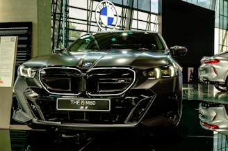 A sleek, modern car is prominently displayed in a showroom with a reflective floor. It has a dark finish and detailed, stylish headlamps. The car's badge is visible on the front grille, and there is a sign with model information next to it.