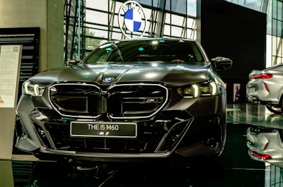 A sleek, modern car is prominently displayed in a showroom with a reflective floor. It has a dark finish and detailed, stylish headlamps. The car's badge is visible on the front grille, and there is a sign with model information next to it.