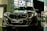 A sleek, modern car is prominently displayed in a showroom with a reflective floor. It has a dark finish and detailed, stylish headlamps. The car's badge is visible on the front grille, and there is a sign with model information next to it.