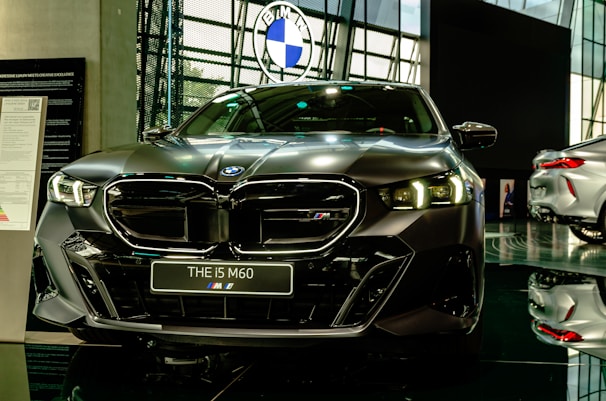 A sleek, modern car is prominently displayed in a showroom with a reflective floor. It has a dark finish and detailed, stylish headlamps. The car's badge is visible on the front grille, and there is a sign with model information next to it.