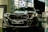 A sleek, modern car is prominently displayed in a showroom with a reflective floor. It has a dark finish and detailed, stylish headlamps. The car's badge is visible on the front grille, and there is a sign with model information next to it.