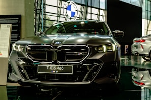 A sleek, modern car is prominently displayed in a showroom with a reflective floor. It has a dark finish and detailed, stylish headlamps. The car's badge is visible on the front grille, and there is a sign with model information next to it.