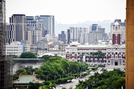 The image features a cityscape with a blend of modern and traditional architecture. In the foreground, lush green trees line a wide street. The midground showcases a prominent historical building with red and white detailing, possibly a government or historical structure. In the background, tall contemporary buildings dominate the skyline.