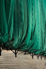 A large green net or fabric is draped over a series of black metal supports attached to a beige corrugated wall. The net hangs in loose folds, partially covering the supports and casting shadows on the wall.