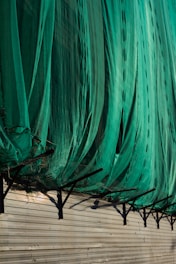 A large green net or fabric is draped over a series of black metal supports attached to a beige corrugated wall. The net hangs in loose folds, partially covering the supports and casting shadows on the wall.