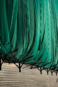 A large green net or fabric is draped over a series of black metal supports attached to a beige corrugated wall. The net hangs in loose folds, partially covering the supports and casting shadows on the wall.