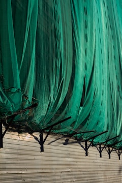 A large green net or fabric is draped over a series of black metal supports attached to a beige corrugated wall. The net hangs in loose folds, partially covering the supports and casting shadows on the wall.