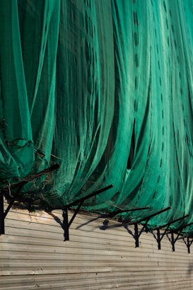 A large green net or fabric is draped over a series of black metal supports attached to a beige corrugated wall. The net hangs in loose folds, partially covering the supports and casting shadows on the wall.
