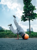 A person wearing a bright orange helmet is performing a breakdancing move on an asphalt road, with one hand on the ground and legs in the air. The surroundings include green grass, trees, and a few buildings in the distance under a partly cloudy blue sky.