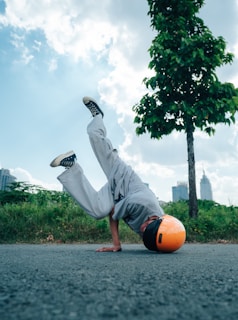 A person wearing a bright orange helmet is performing a breakdancing move on an asphalt road, with one hand on the ground and legs in the air. The surroundings include green grass, trees, and a few buildings in the distance under a partly cloudy blue sky.