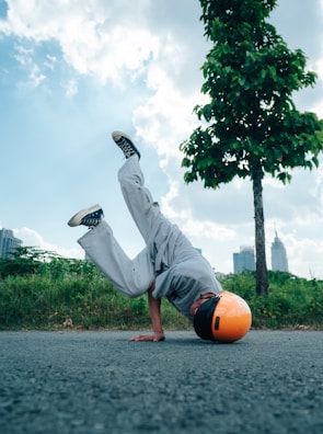 A person wearing a bright orange helmet is performing a breakdancing move on an asphalt road, with one hand on the ground and legs in the air. The surroundings include green grass, trees, and a few buildings in the distance under a partly cloudy blue sky.