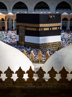 A large crowd gathers around the Kaaba in the center of the Grand Mosque in Mecca. The Kaaba is covered with a black and gold embroidered cloth. The surrounding mosque is filled with arches and intricate architectural elements. People are dressed in various attire, many holding umbrellas to shield from the sun.