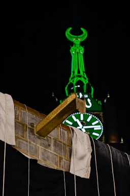 A serene evening view of the Kaaba illuminated under a deep emerald sky with subtle gold geometric patterns framing the scene.