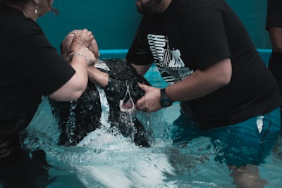 Two people assist another person being submerged in water, possibly for a religious ceremony like baptism. The setting is a shallow pool with turquoise water, and all individuals are wearing dark clothing.
