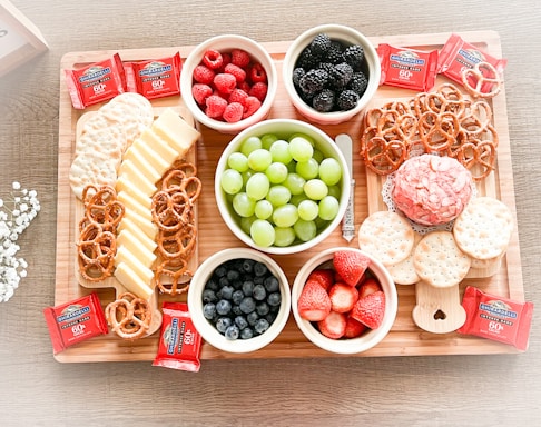 On a wooden tray, there's an assortment of snacks including bowls of raspberries, blackberries, blueberries, strawberries, and green grapes. Slices of cheese and pretzels are arranged neatly alongside round crackers. There are also several squares of Ghirardelli chocolate, and a decorative cheese ball is surrounded by more crackers.
