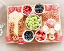 On a wooden tray, there's an assortment of snacks including bowls of raspberries, blackberries, blueberries, strawberries, and green grapes. Slices of cheese and pretzels are arranged neatly alongside round crackers. There are also several squares of Ghirardelli chocolate, and a decorative cheese ball is surrounded by more crackers.
