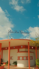 A retro-styled beach resort entrance with a circular roof supported by coral-colored pillars. The name 'Lido San Giovanni' is displayed in cursive on top. The sky is bright with a few scattered clouds, and there are some small plants near the building.