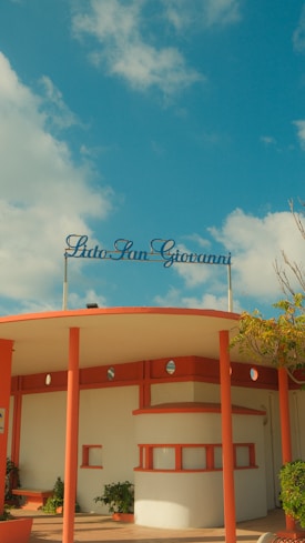 A retro-styled beach resort entrance with a circular roof supported by coral-colored pillars. The name 'Lido San Giovanni' is displayed in cursive on top. The sky is bright with a few scattered clouds, and there are some small plants near the building.