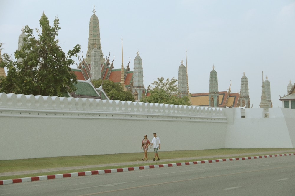 two people walking down a street past a white wall