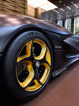 A sleek, modern car with a focus on its distinctive yellow and black alloy wheel, featuring a Michelin tire. The body of the car has a matte black finish, displayed in a well-lit, contemporary showroom with a wooden floor and large windows.