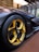 A sleek, modern car with a focus on its distinctive yellow and black alloy wheel, featuring a Michelin tire. The body of the car has a matte black finish, displayed in a well-lit, contemporary showroom with a wooden floor and large windows.