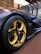 A sleek, modern car with a focus on its distinctive yellow and black alloy wheel, featuring a Michelin tire. The body of the car has a matte black finish, displayed in a well-lit, contemporary showroom with a wooden floor and large windows.