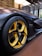 A sleek, modern car with a focus on its distinctive yellow and black alloy wheel, featuring a Michelin tire. The body of the car has a matte black finish, displayed in a well-lit, contemporary showroom with a wooden floor and large windows.