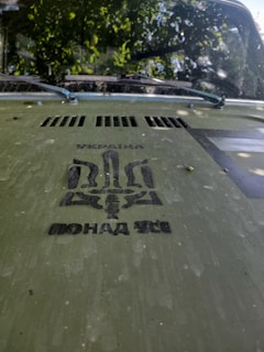 The hood of a vehicle displays a stencil design in black ink. The design includes text written in Cyrillic, along with a stylized trident symbol. The hood is a greenish color and appears slightly worn with spots. The vehicle’s windshield is visible, and there is a reflection of leaves and branches above it, indicating the vehicle is parked under a tree.