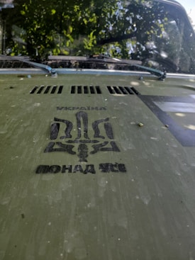 The hood of a vehicle displays a stencil design in black ink. The design includes text written in Cyrillic, along with a stylized trident symbol. The hood is a greenish color and appears slightly worn with spots. The vehicle&rsquo;s windshield is visible, and there is a reflection of leaves and branches above it, indicating the vehicle is parked under a tree.