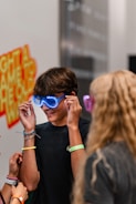 A person wearing glowing blue novelty glasses, surrounded by friends. The scene appears to be indoors, possibly at an event or party with a vibrant and playful atmosphere. A wall in the background has colorful text and designs.