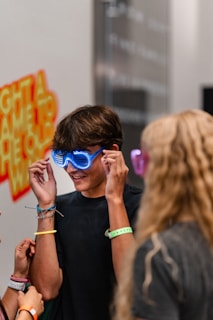 A person wearing glowing blue novelty glasses, surrounded by friends. The scene appears to be indoors, possibly at an event or party with a vibrant and playful atmosphere. A wall in the background has colorful text and designs.