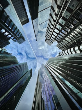 A dramatic upward perspective of modern skyscrapers converging towards the center, showcasing the reflective glass surfaces and metallic structures against a partly cloudy sky.