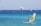 Windsurfers and a kite surfer navigate the vibrant blue sea with visible wind gusts. The ocean rests against a backdrop of distant, hazy mountains, while the sky above is clear with a hint of haze.