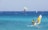 Windsurfers and a kite surfer navigate the vibrant blue sea with visible wind gusts. The ocean rests against a backdrop of distant, hazy mountains, while the sky above is clear with a hint of haze.