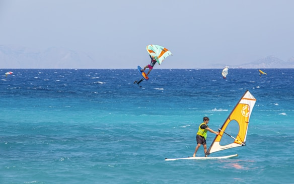 Windsurfers and a kite surfer navigate the vibrant blue sea with visible wind gusts. The ocean rests against a backdrop of distant, hazy mountains, while the sky above is clear with a hint of haze.