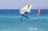 Two individuals are engaging in water sports in a vibrant blue sea under a clear sky. One person is riding a foil board with an inflatable wing, caught mid-air, showcasing skill and balance. The other person is windsurfing, gliding smoothly over the water. The scene conveys action and adventure along a scenic coastal backdrop.