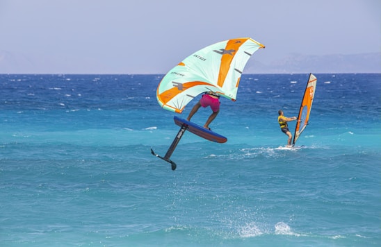 Two individuals are engaging in water sports in a vibrant blue sea under a clear sky. One person is riding a foil board with an inflatable wing, caught mid-air, showcasing skill and balance. The other person is windsurfing, gliding smoothly over the water. The scene conveys action and adventure along a scenic coastal backdrop.