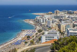 A coastal cityscape with a vibrant blue sea in the background, lined with numerous white and colorful buildings. A beach with sun loungers and umbrellas stretches along the shore, while a road runs parallel to the coastline. The scene includes a parasail in the sky and a few boats in the water.
