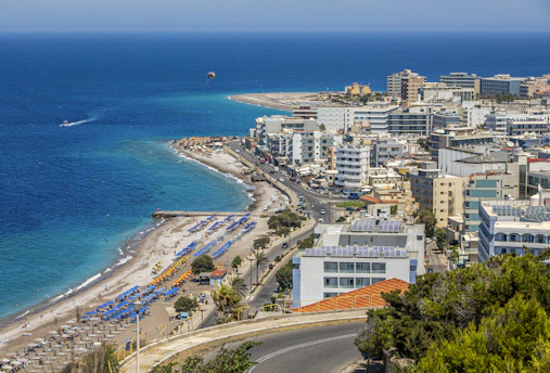 A coastal cityscape with a vibrant blue sea in the background, lined with numerous white and colorful buildings. A beach with sun loungers and umbrellas stretches along the shore, while a road runs parallel to the coastline. The scene includes a parasail in the sky and a few boats in the water.