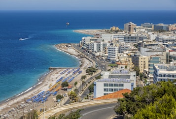 A coastal cityscape with a vibrant blue sea in the background, lined with numerous white and colorful buildings. A beach with sun loungers and umbrellas stretches along the shore, while a road runs parallel to the coastline. The scene includes a parasail in the sky and a few boats in the water.
