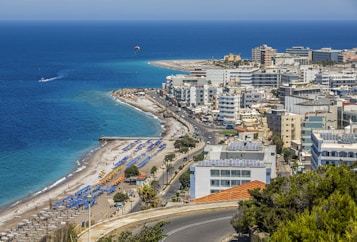 A coastal cityscape with a vibrant blue sea in the background, lined with numerous white and colorful buildings. A beach with sun loungers and umbrellas stretches along the shore, while a road runs parallel to the coastline. The scene includes a parasail in the sky and a few boats in the water.