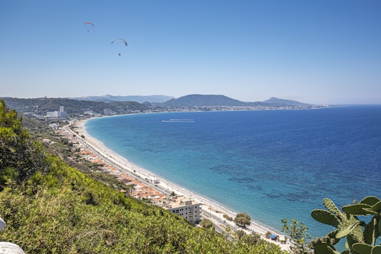 A scenic coastal landscape with a winding shoreline, lush green foliage in the foreground, and a vast expanse of blue sea under a clear sky. Two paragliders are visible in the sky, adding a dynamic element to the tranquil scene. Buildings line the shore, and hills provide a backdrop on the horizon.