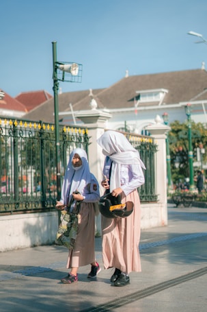Two girls wearing school uniforms and headscarves are walking on a sunlit street. One carries a helmet and a phone, the other holds a patterned bag. A decorative fence and a historic building form the background.