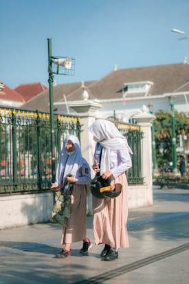 Two girls wearing school uniforms and headscarves are walking on a sunlit street. One carries a helmet and a phone, the other holds a patterned bag. A decorative fence and a historic building form the background.
