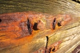 Weathered wooden boards with rusted metal bolts. The wood has visible grains and textures with varying shades of brown and green. Rust stains from the bolts have spread onto the surrounding wood, creating a mix of orange and red hues.