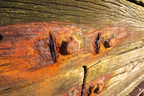 Weathered wooden boards with rusted metal bolts. The wood has visible grains and textures with varying shades of brown and green. Rust stains from the bolts have spread onto the surrounding wood, creating a mix of orange and red hues.